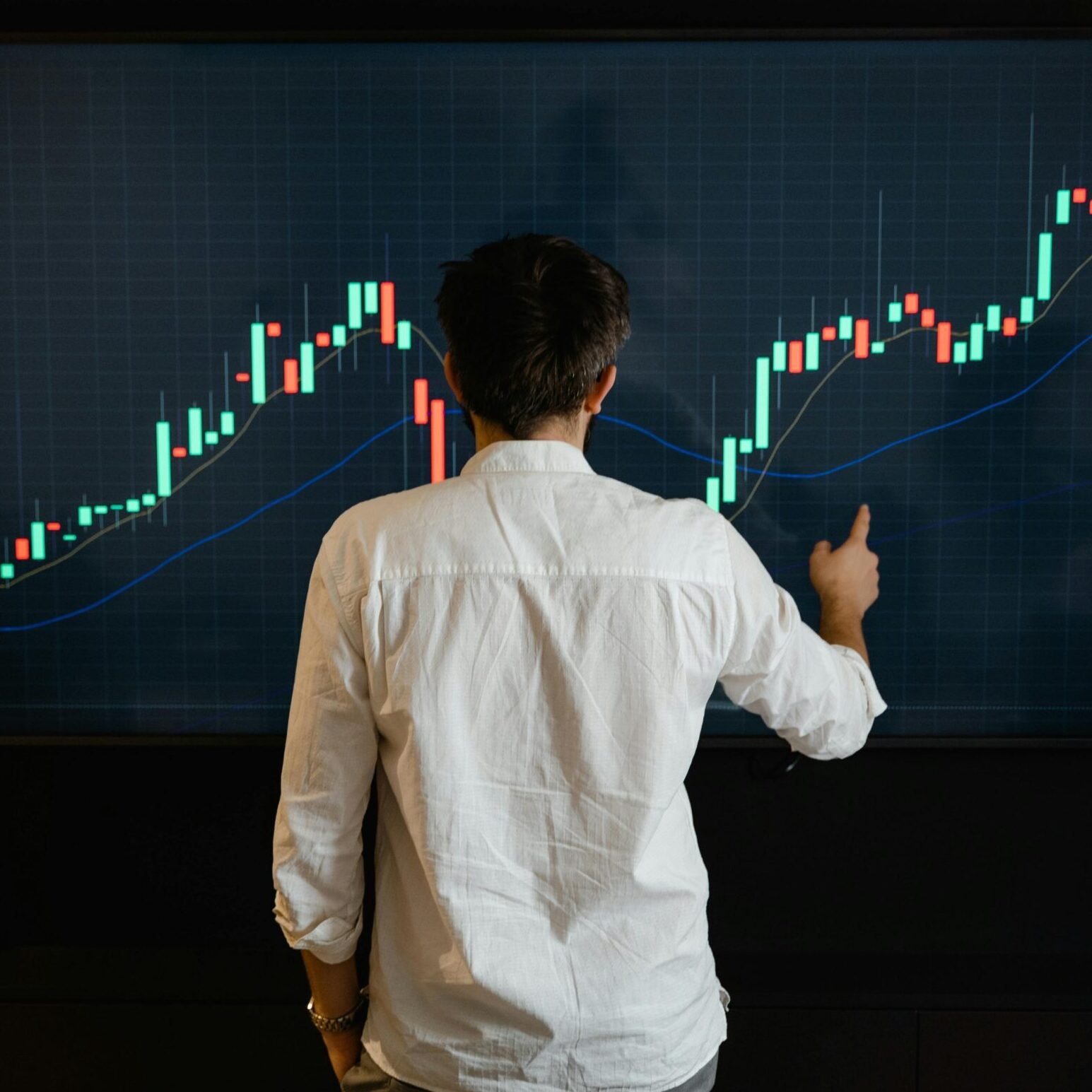 Adult male reviewing stock market data on a large display screen indoors.